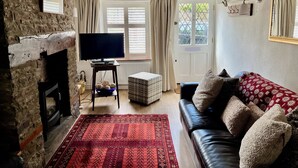 A cozy living room with a stone fireplace, a flat-screen TV, a red patterned rug, a black leather sofa, and a door leading outside. - Pretty cottage, perfect readers' retreat in centre of Hay, parking & fast wifi (Hay-on-Wye)