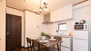Een kleine keuken met witte kasten, een magnetron en een koelkast. Er staat een eettafel met stoelen en een potplant.
