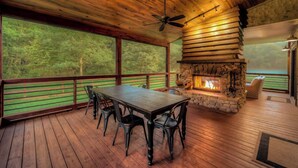 A wooden deck with a stone fireplace, a dining table with chairs, and a view of the forest. - Firefly Farmhouse - Riverfront | Modern | 2 Bedroom w loft (Blue Ridge)