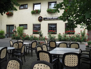 Un espace extérieur aménagé avec des chaises et des tables en osier, un bâtiment orné de jardinières et une enseigne indiquant la présence d’un restaurant.