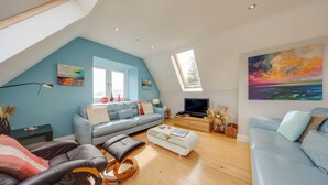 A modern living room with a sloped ceiling, a blue sofa, a white ottoman, and a flat-screen TV.