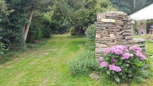 Un chemin de pierres mène à une construction en pierre sur laquelle est apposée une plaque, le tout étant entouré d’une végétation luxuriante et d’une plante en fleurs.