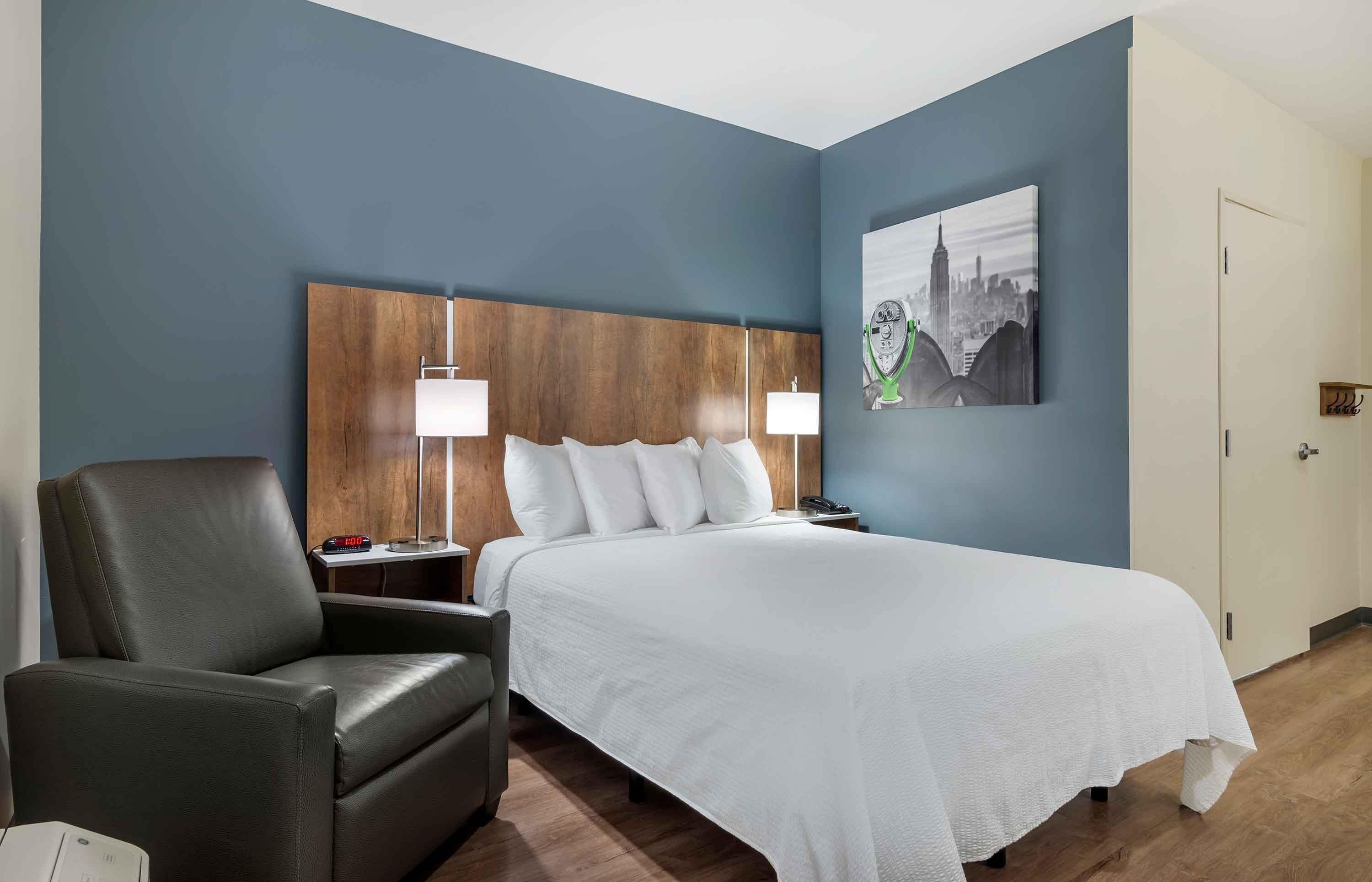 Una habitación de hotel con una cama, una silla, una mesita de noche, una lámpara y un cuadro con una vista urbana en la pared.