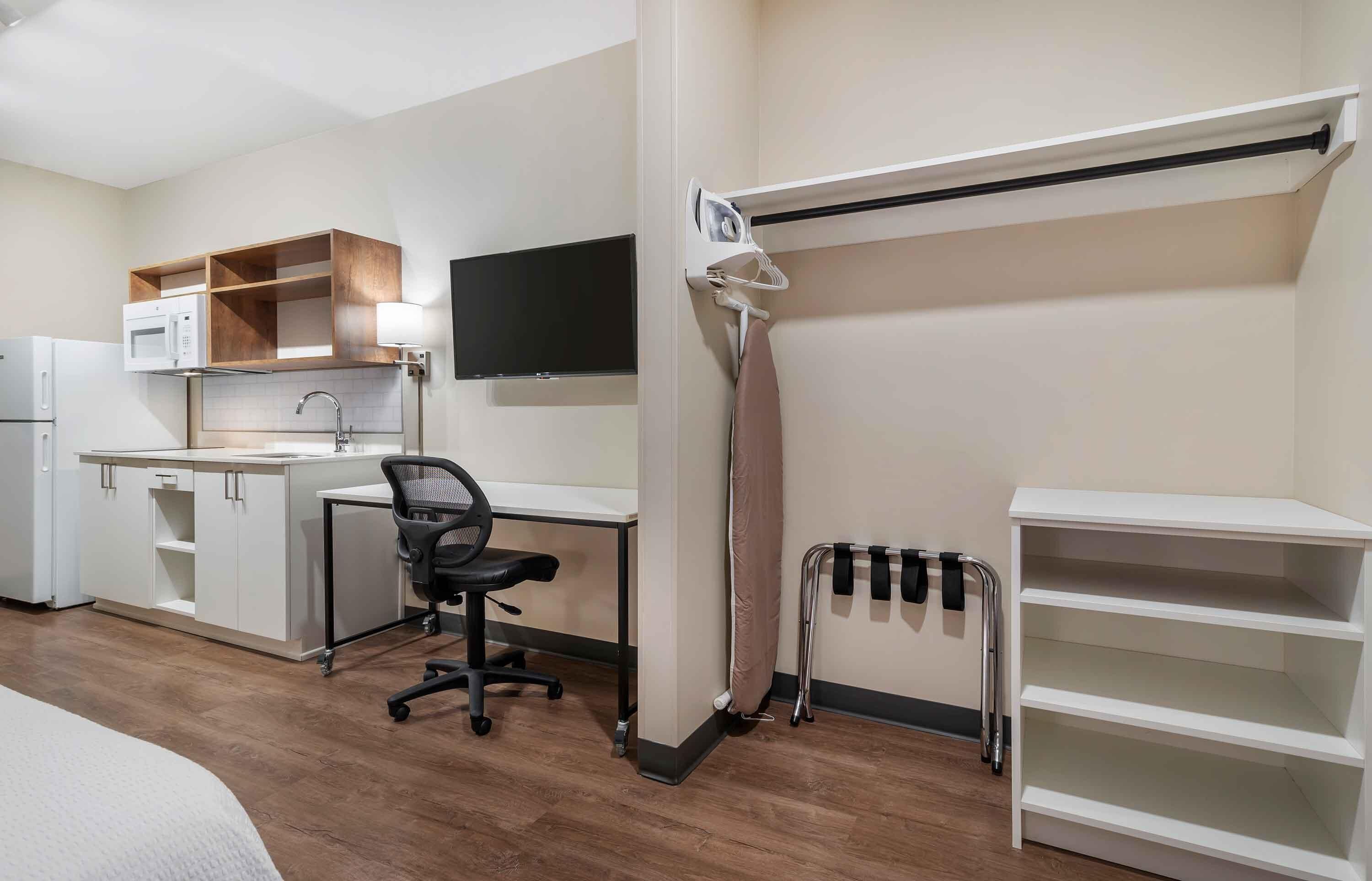 Una habitación de hotel compacta con kitchenette, un escritorio y una silla, un televisor montado en la pared y una cama con cabecera blanca.