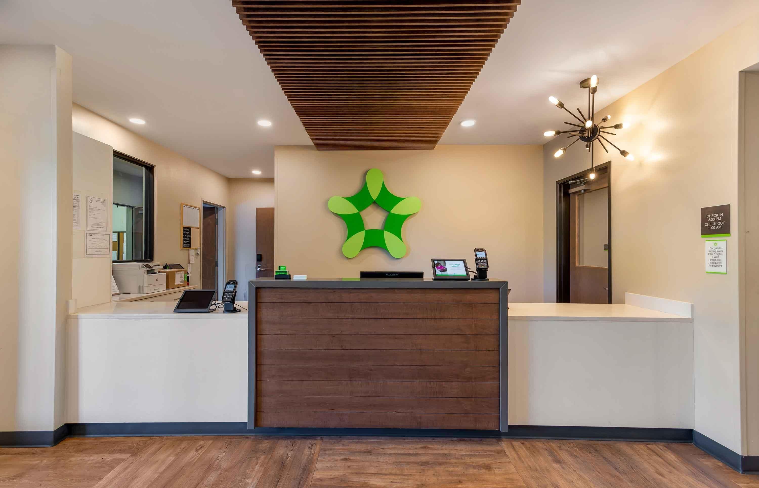 A reception desk with a wooden panel, a green logo, a digital screen, and a check-in kiosk.
