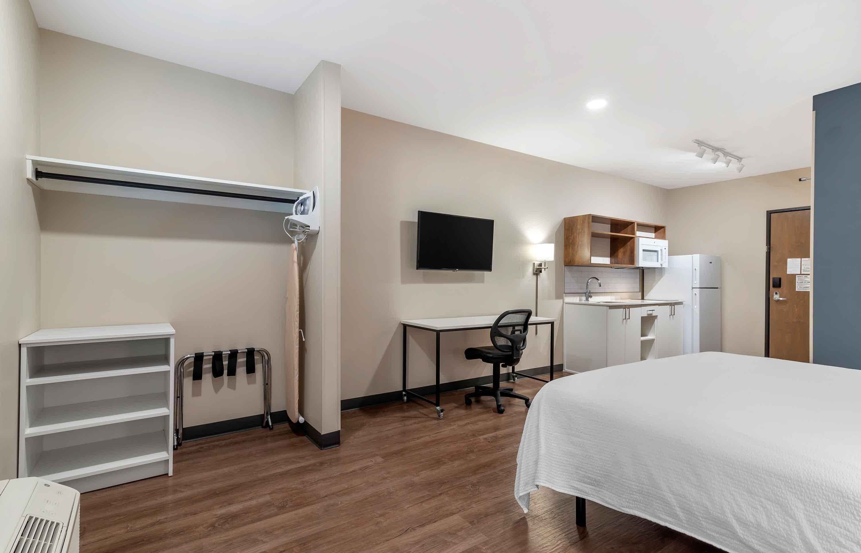 A hotel room with a bed, a desk with a chair, a TV, a kitchenette, and a wardrobe.