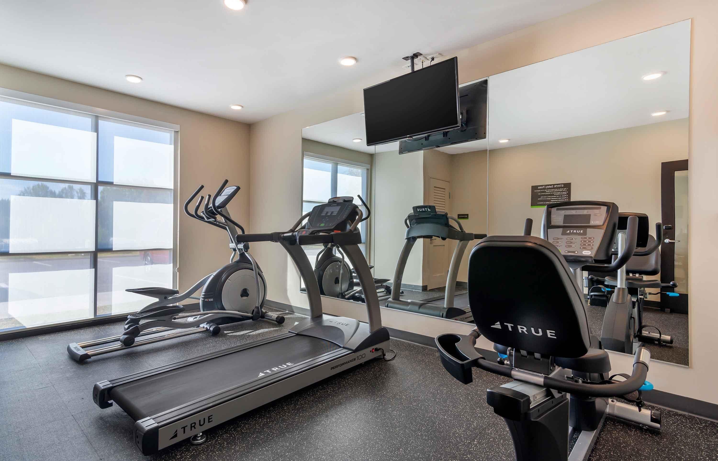 Une salle de sport équipée de tapis de course, d’un vélo elliptique et d’un vélo d’appartement, avec un grand miroir et un téléviseur fixé au mur.