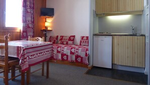 Una habitación pequeña y acogedora con una mesa de madera, una cama con ropa de cama de diseño rojo y blanco, un televisor pequeño montado en la pared y una cocina compacta con armarios de madera y un lavamanos.