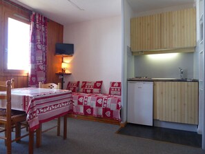 A small, cozy room with a wooden table, a bed with red and white patterned bedding, a small TV mounted on the wall, and a compact kitchen area with wooden cabinets and a sink.