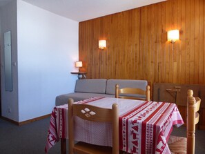 A room with a wooden wall, a sofa, a table with a patterned tablecloth, and two wall-mounted lamps.