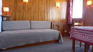 Una habitación con un revestimiento de madera, una cama con cubrecama gris, una pequeña mesa con mantel estampado en rojo y blanco, y dos lámparas fijadas a la pared.