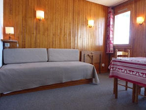 A room with a wooden paneled wall, a bed with a grey bedspread, a small table with a red and white patterned tablecloth, and two wall-mounted lamps.