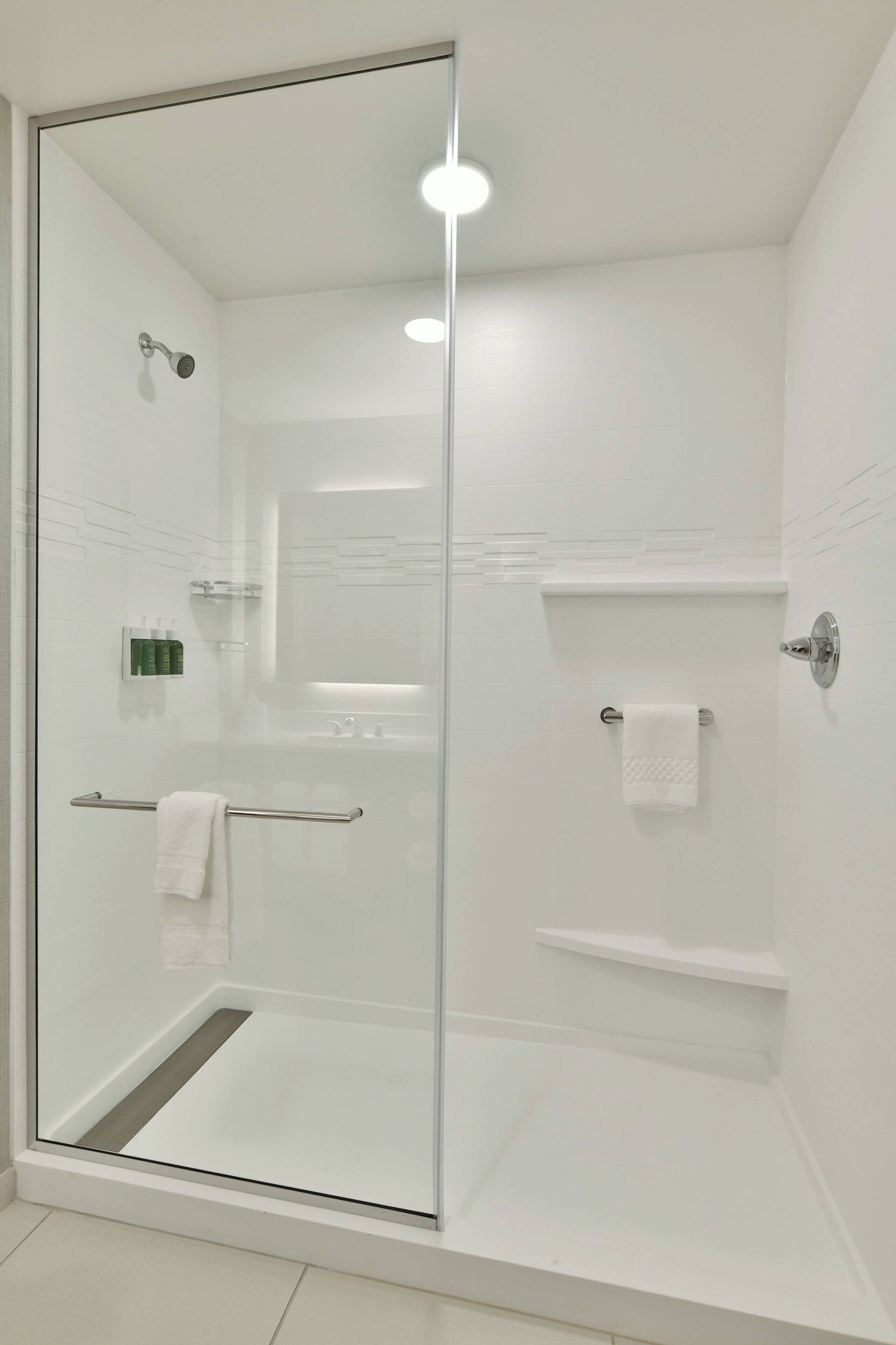A modern shower with a glass door, a white tiled wall, a towel rack, and a shelf with toiletries.