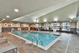 Uma ampla piscina interior com pistas demarcadas, uma cadeira de salva-vidas e uma área de estar com mesas e cadeiras.