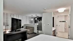 A modern hotel room with a bed, a desk, a TV, a sofa, and a kitchenette.