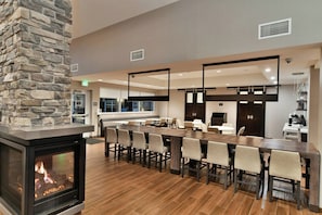 A modern interior with a stone fireplace, a long dining table with chairs, and a kitchen area in the background.