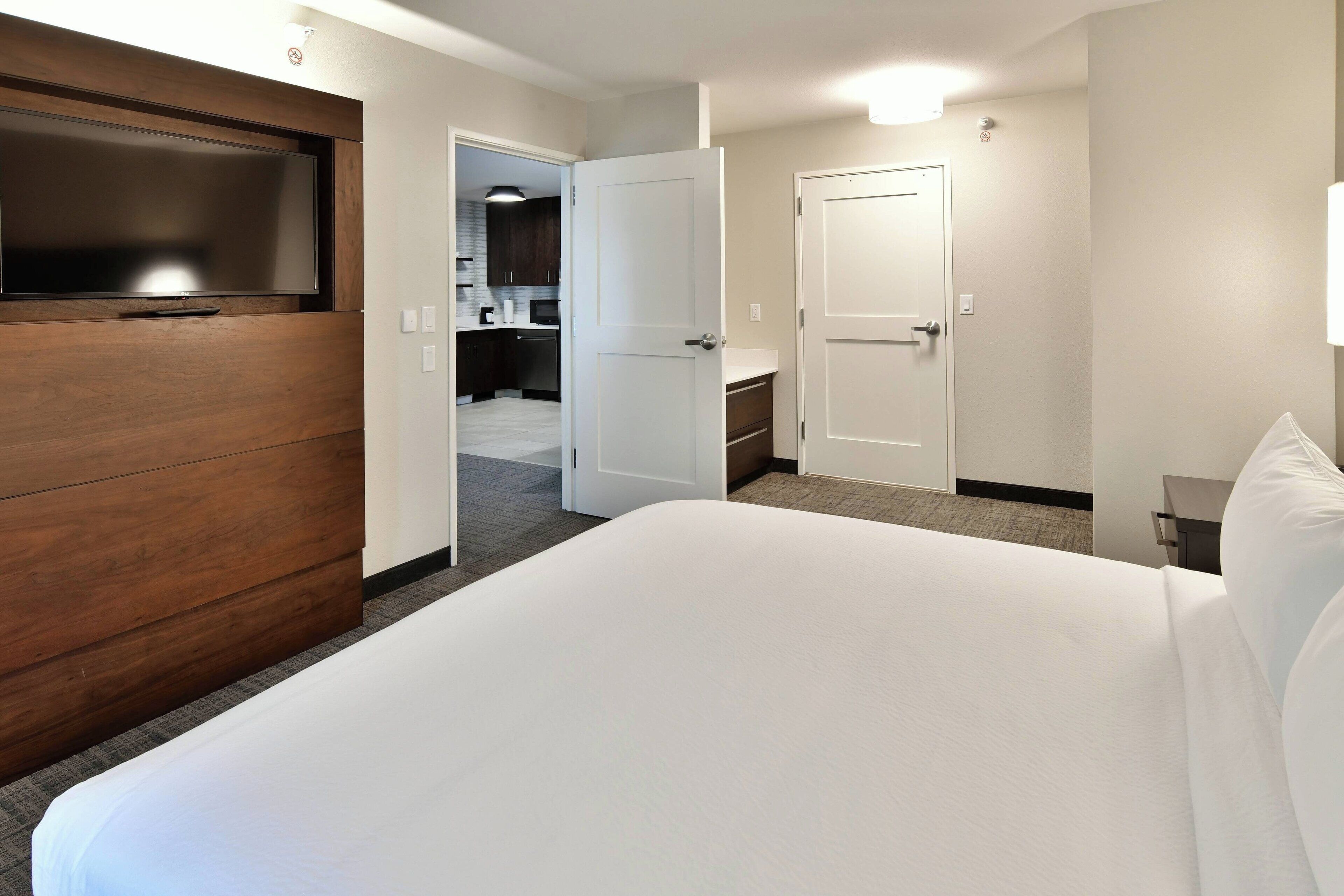 Hotelkamer met een groot bed, een televisie bevestigd aan een houten paneel, een kitchenette op de achtergrond en een badkamerdeur.