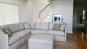Ein modernes Wohnzimmer mit einem grauen Ecksofa, einer Holztreppe und einem gerahmten Bild an der Wand.