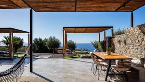 A covered outdoor patio with a wooden table and chairs, a hammock, and a stone wall. - Luxe Coastal Escape with Sea Views - Kumo Villa I (Rethymno)