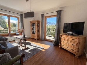 Een woonkamer met een bank, een televisie, een kast en uitzicht op een bergachtig landschap door grote ramen.