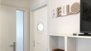 Ein Zimmer mit einer weißen Tür, einer Wanduhr und einem Schild mit der Aufschrift 'HELLO'. Auf Regalen stehen zwei Holzkisten.