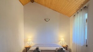 Ein Schlafzimmer mit Holzbalkendecke, einem Bett mit aufgerollten Handtüchern, zwei Nachttischlampen und einem Fenster mit Vorhängen.