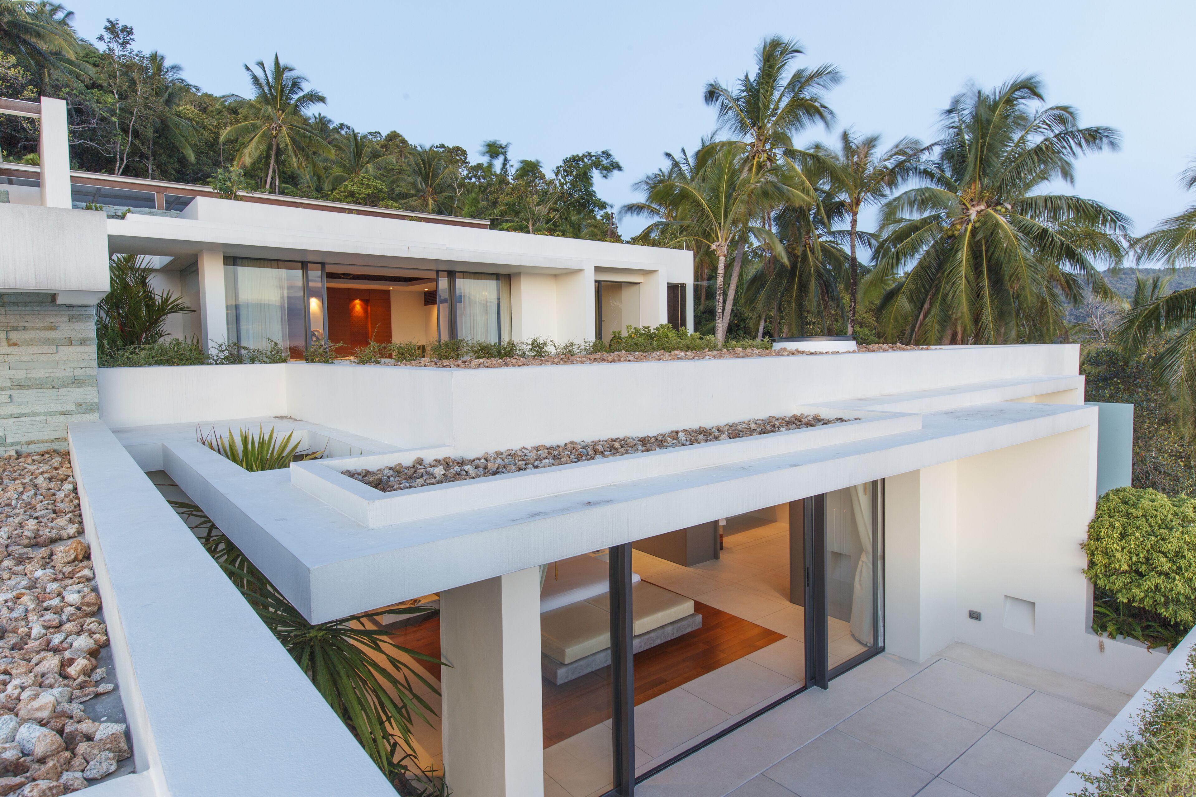 A modern house with a flat roof, large windows, and a balcony overlooking a tropical landscape with palm trees.
