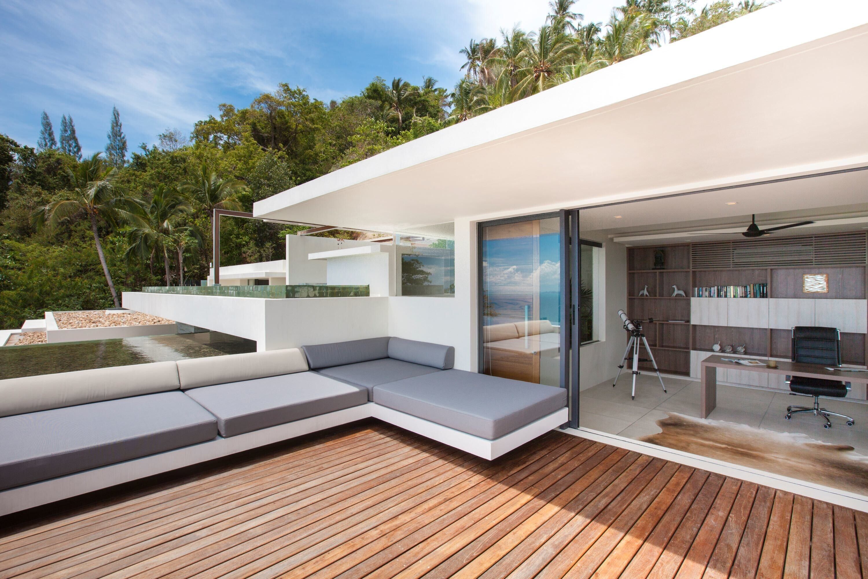 A modern outdoor living area with a wooden deck, built-in seating, and a glass door leading to a room with a desk and bookshelves.