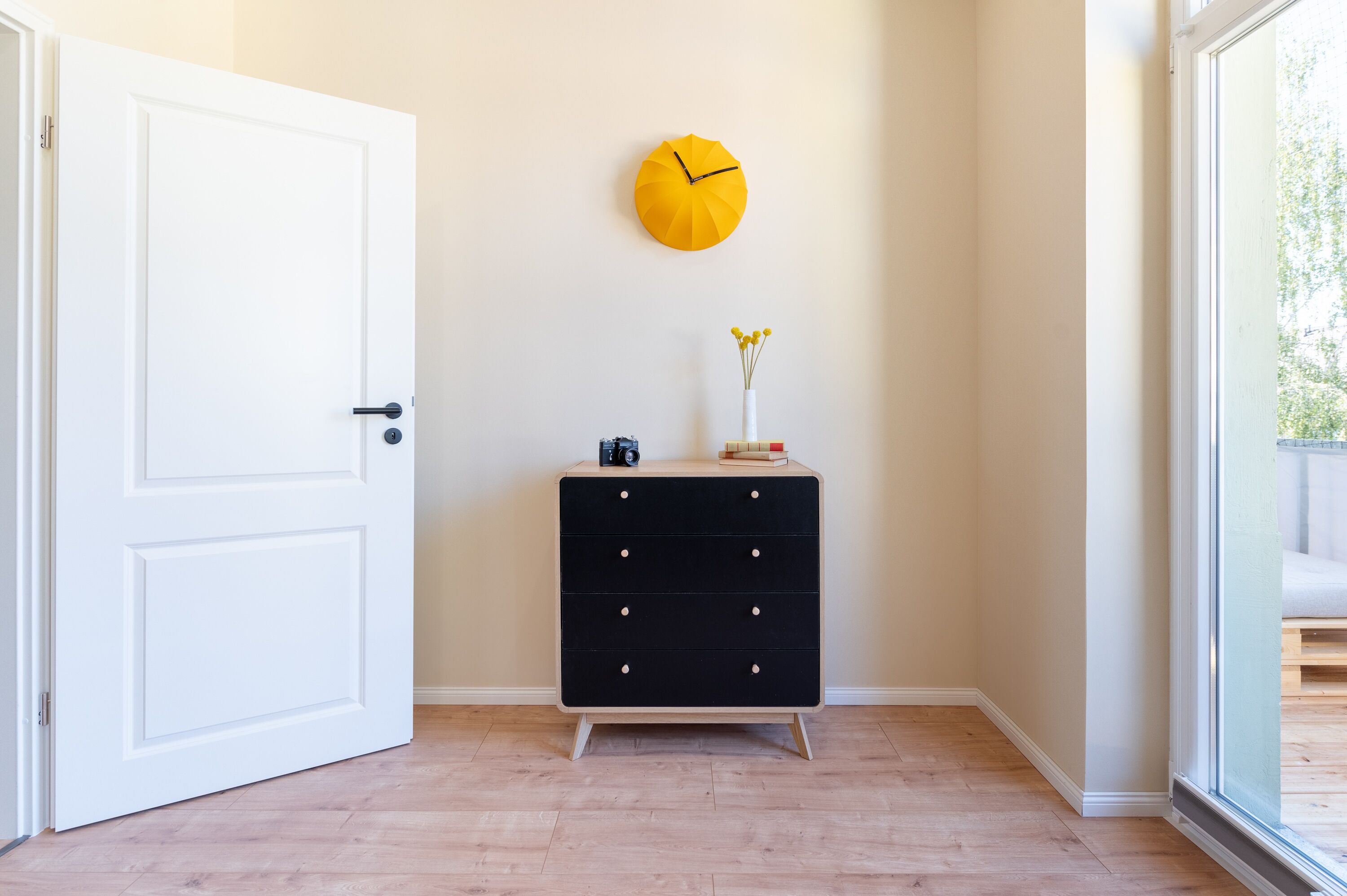 Ein Zimmer mit einer weißen Tür, einer schwarzen Kommode mit Schubladen, einer gelben Wanduhr und einer Vase mit Blumen auf der Kommode.