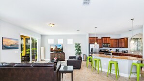 Uma sala de estar moderna com um sofá de couro marrom, uma área de jantar com banquetas verdes e uma cozinha com armários de madeira e eletrodomésticos de aço inoxidável.