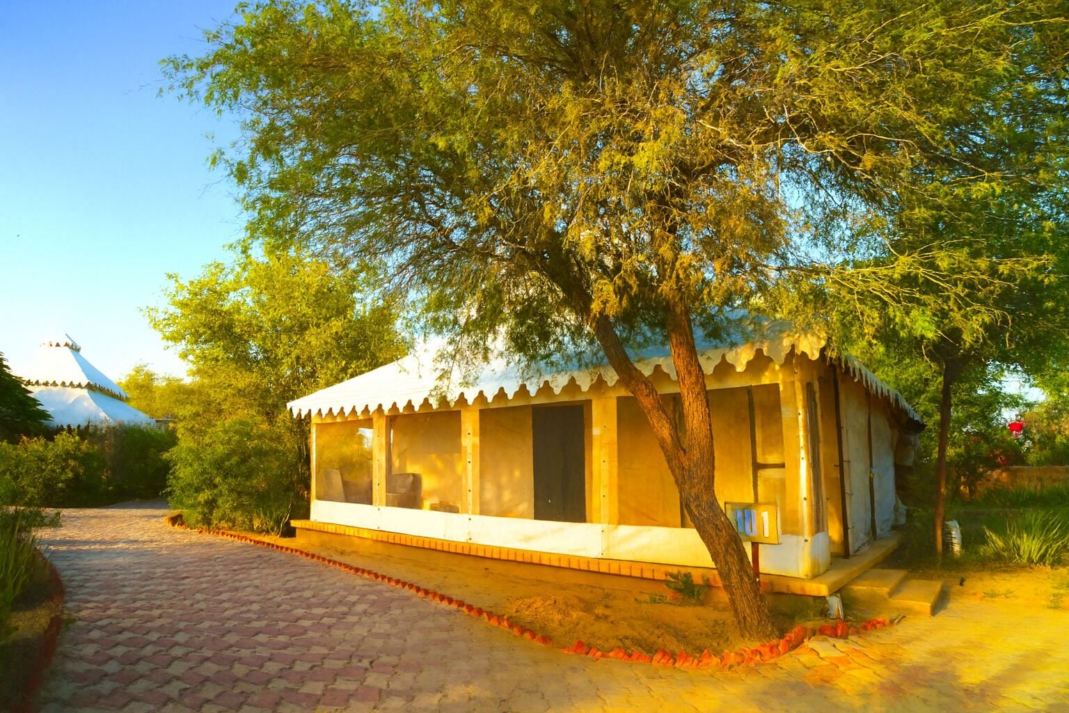 A small, single-story building with a white exterior and a thatched roof, surrounded by trees and a paved path.