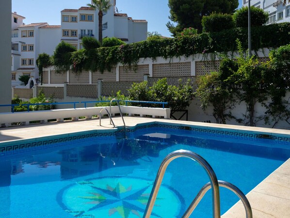 A swimming pool with a geometric tile pattern, surrounded by a white wall with greenery and a palm tree.