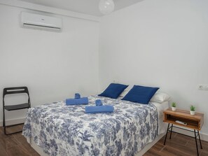Ein Schlafzimmer mit einem Bett, einem Sessel, einem Nachttisch mit Pflanze und einer Klimaanlage an der Wand.