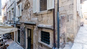 Un edificio de piedra con varias ventanas, algunas con contraventanas, y una puerta. Hay mesas y sillas al aire libre, lo que sugiere un café o restaurante.