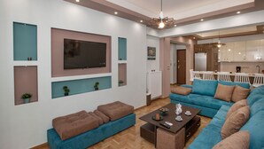 A modern living room with a large TV, blue sofas, a coffee table, and a kitchen area in the background. - Stunning Apt near Acropolis Museum by GHH (Athens)