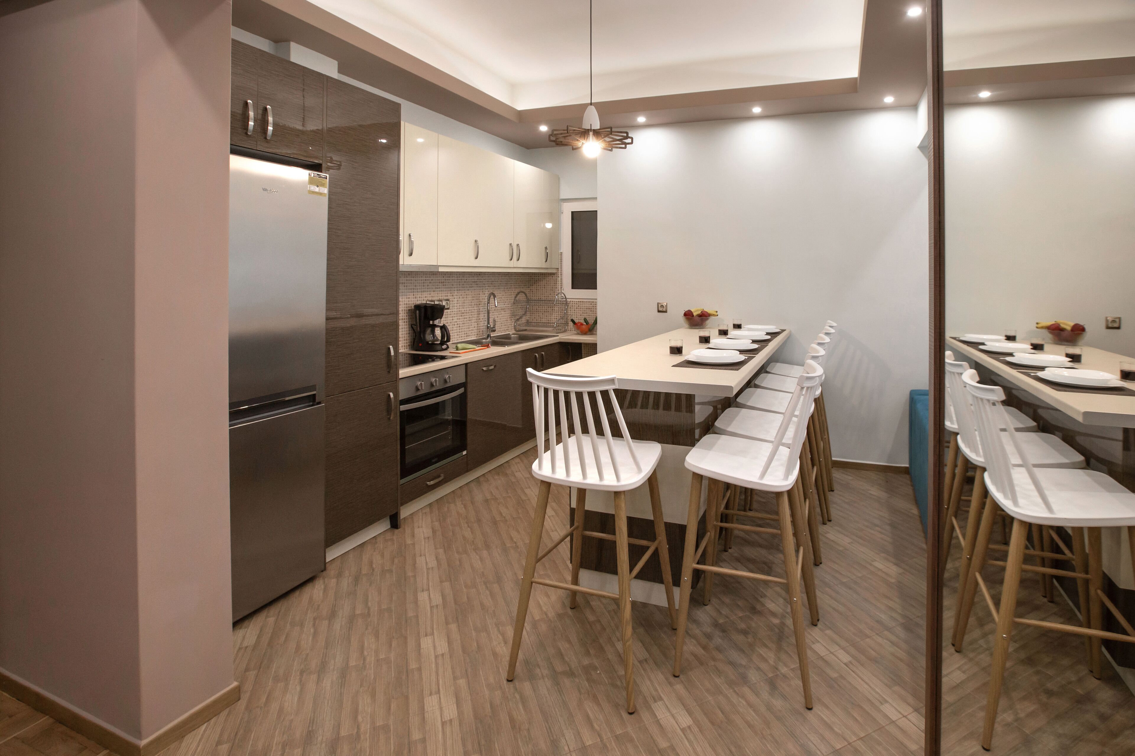 A modern kitchen with a built-in refrigerator, oven, and coffee maker. There are white bar stools and a long dining table with plates and glasses set.