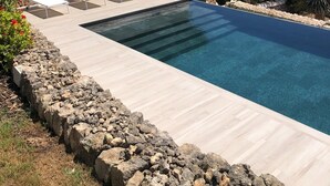 Una piscina con sillones de descanso, una pared de piedra y vistas al mar y edificios.