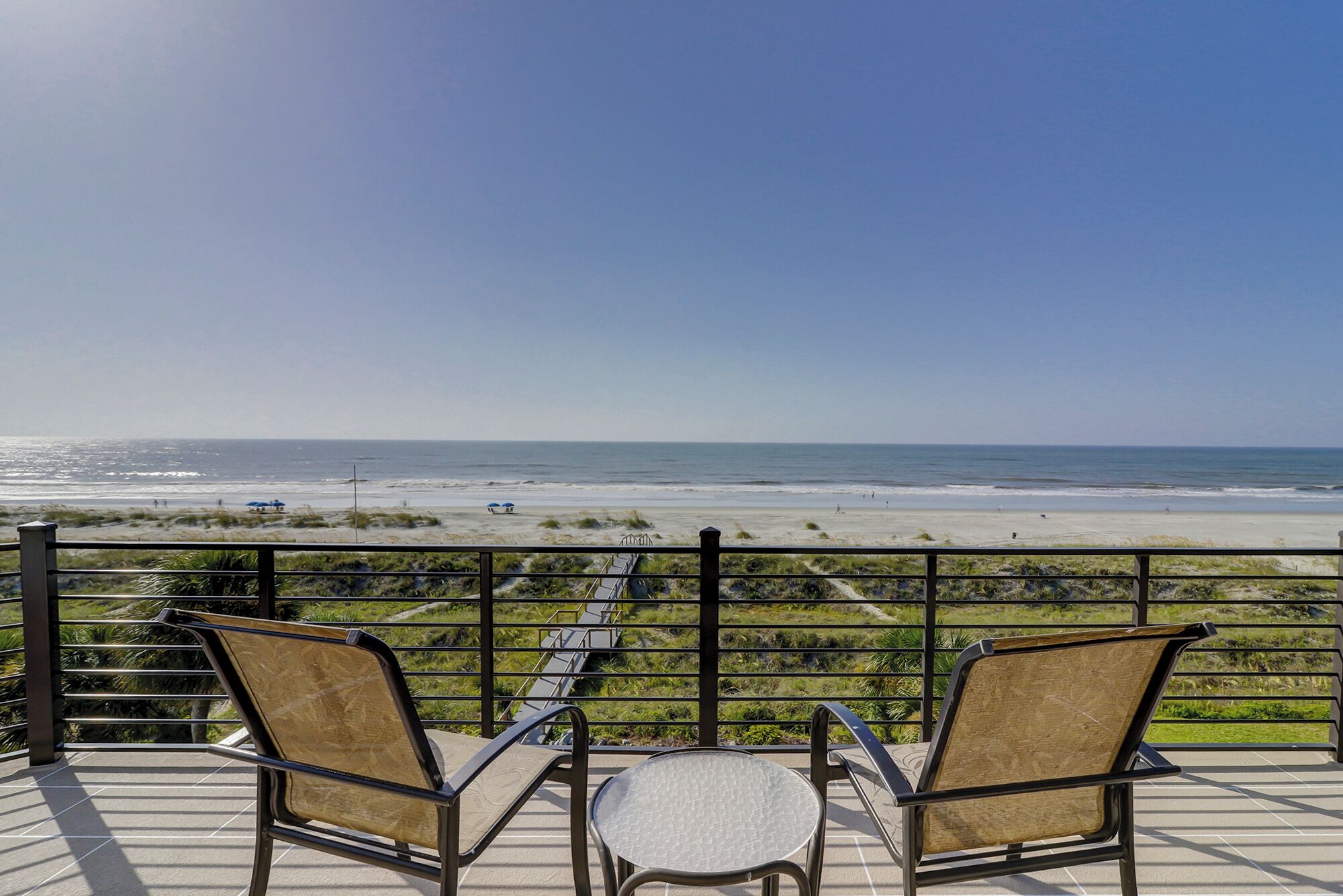 A balcony with two chairs and a small table, overlooking a beach with a clear view of the ocean.