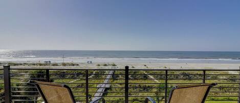 A balcony with two chairs and a small table, overlooking a beach with a clear view of the ocean.