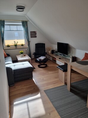 Ein modernes Loft mit hohen Decken, einem großen Fenster mit Vorhängen, einem schwarzen Bürostuhl, einem Holzesstisch, einem Fernseher und einem Sofa.