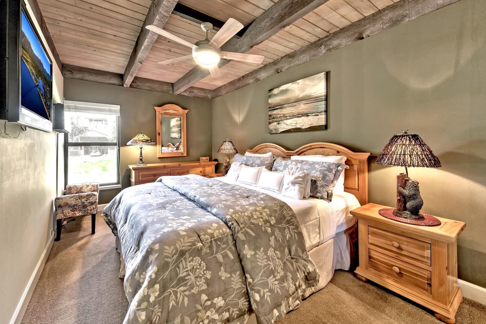 Une chambre avec un grand lit, une tête de lit en bois, un ventilateur de plafond, une table de chevet avec une lampe et une fenêtre donnant sur l’extérieur.
