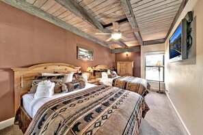 Ein Schlafzimmer mit zwei Betten, Holz-Kopfteilen, einem Deckenventilator, einem an der Wand montierten Fernseher und einem Fenster mit Blick ins Freie.