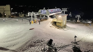 Ein Skigebiet bei Nacht mit einem kleinen Gebäude, einem überdachten Bereich und einer Seilbahn im Hintergrund.