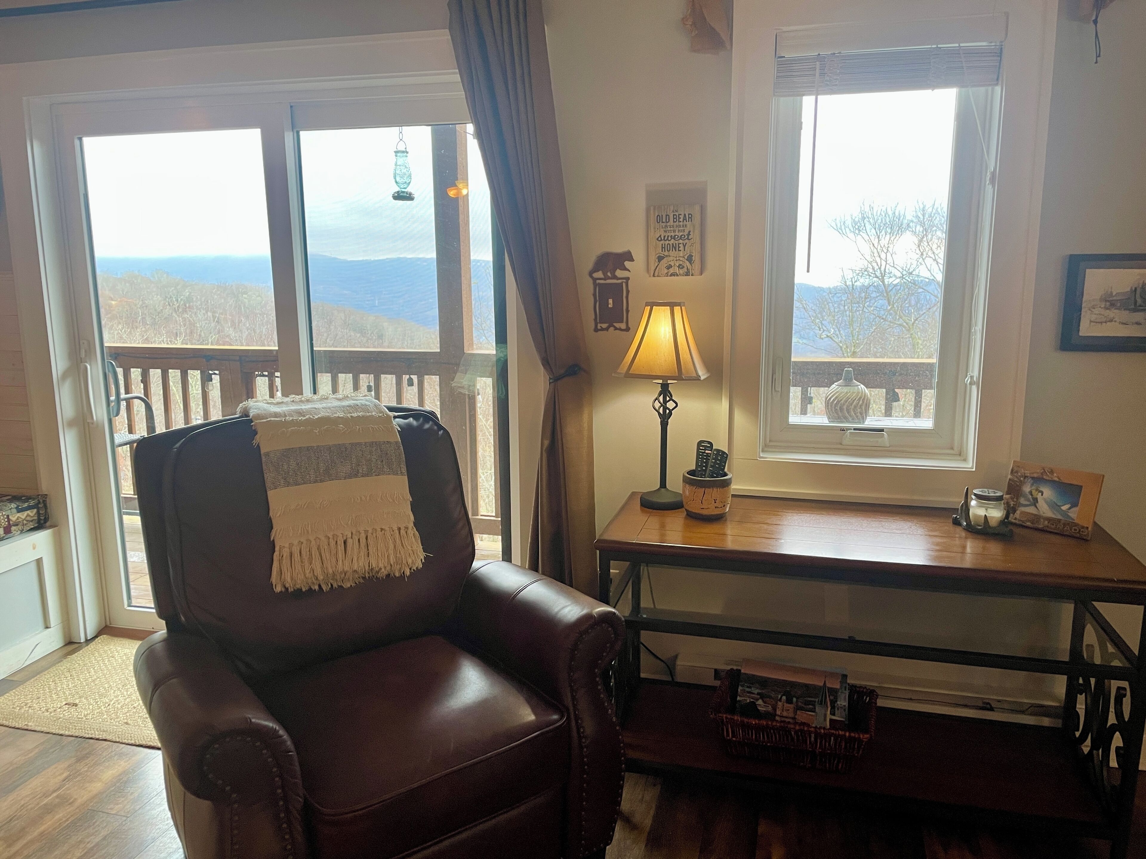 A cozy living room with a leather armchair, a wooden table with a lamp, a framed picture, and a view of the outdoors through large windows.