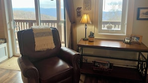 Ein gemütliches Wohnzimmer mit Ledersessel, Holztisch mit Lampe, gerahmtem Bild und Blick ins Freie durch große Fenster.