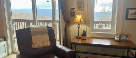 A cozy living room with a leather armchair, a wooden table with a lamp, a framed picture, and a view of the outdoors through large windows.