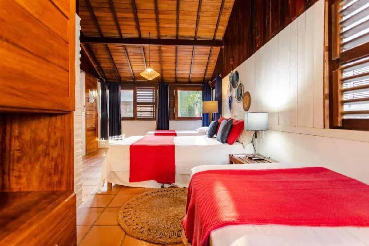 Ein Zimmer mit zwei Betten, Holzbalkendecke, einem großen Fenster und einer gewebten Matte auf dem Boden.
