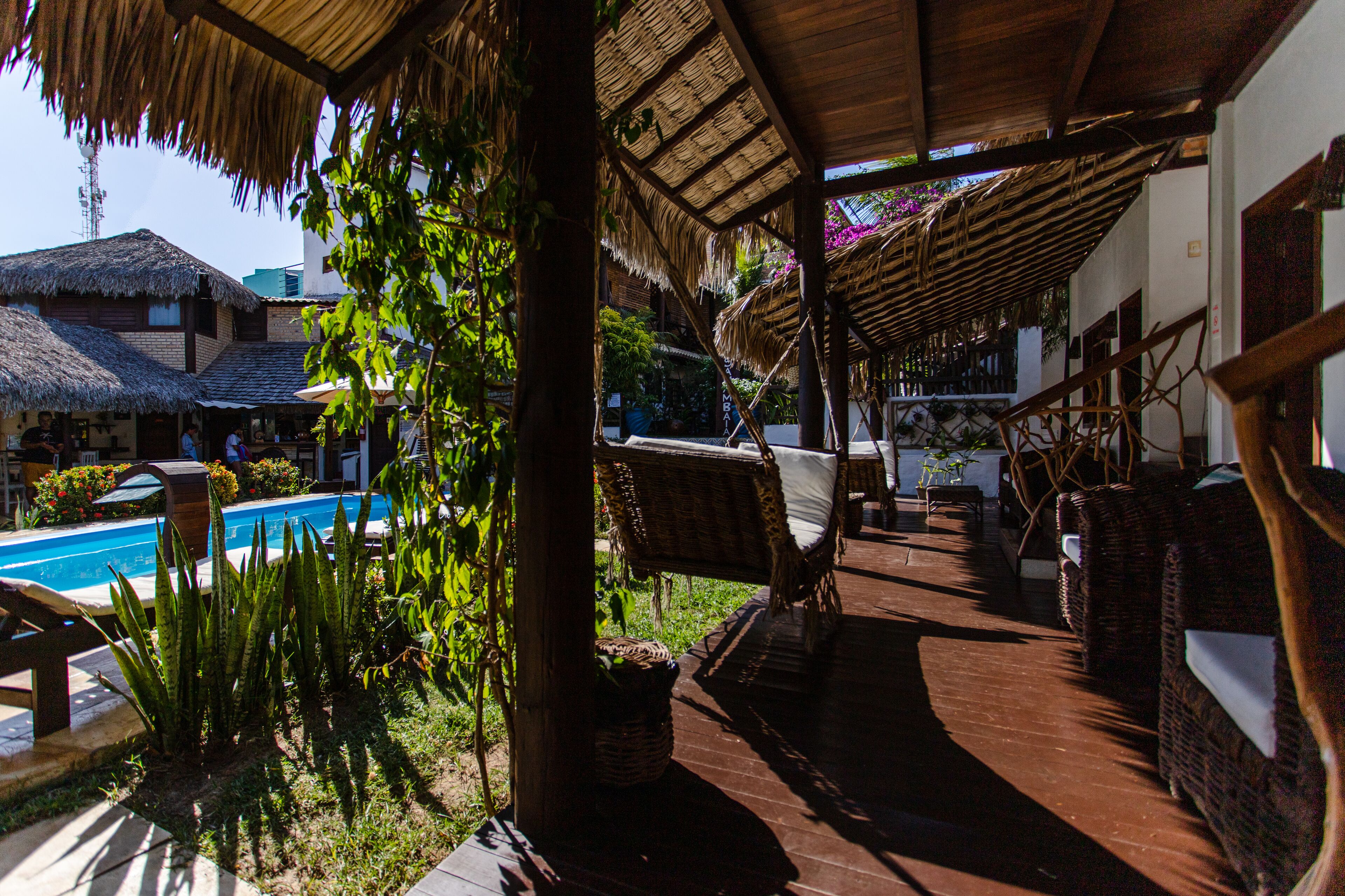 Una terraza de madera con mobiliario de mimbre, una piscina y una estructura con techo de paja.