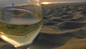 Una copa de vino blanco con condensación, sobre una playa con dunas de arena y el sol al fondo.