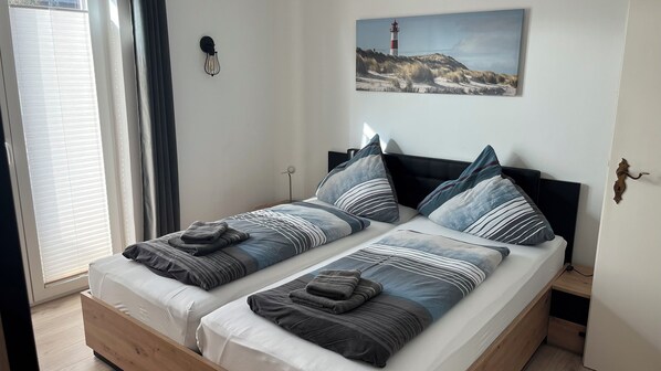 A double bed with striped bedding, a nightstand, a lamp, a painting of a lighthouse, and a window with blinds.